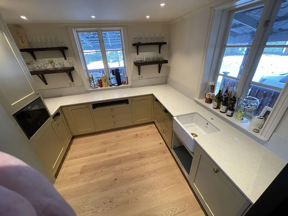 Köksbänkskiva i vit Silestone Snowy Ibiza, polerad, i ett U-format kök med salviegröna shakerluckor och ekgolv i Enköping.