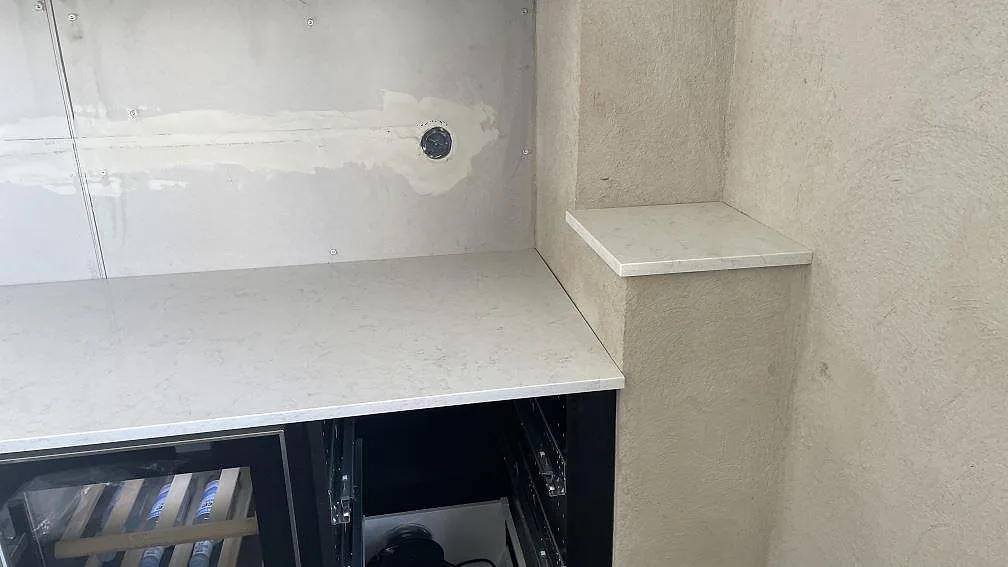 L-formad köksbänk i vit Silestone Snowy Ibiza med hörnhylla i samma sten över vinkylarna i ett kök under montering i Glumslöv.