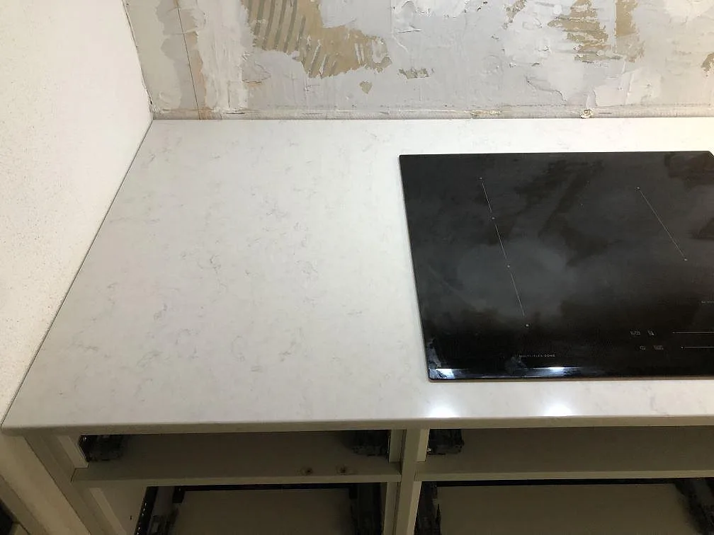 Polerad köksbänkskiva i vit Silestone Snowy Ibiza med infälld svart induktionshäll mot oputsad vägg i ett kök i Göteborg.
