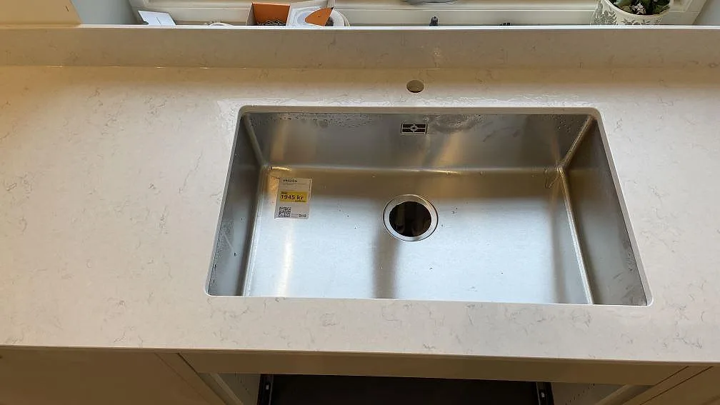 Rostfri diskho i en polerad Silestone Snowy Ibiza-bänkskiva med diskret grå ådring och stänkskydd vid fönstret i ett kök i Gränna.