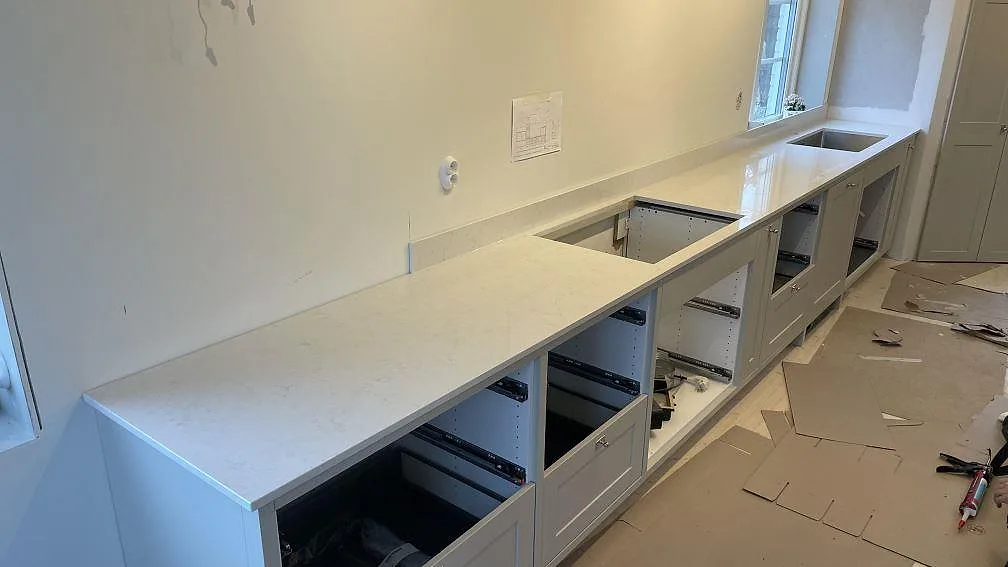 Köksbänkskiva i vit polerad Silestone Snowy Ibiza med 12 mm stänkskydd, rostfri diskho och hällurtag i en lång köksrad under montering i Gränna.