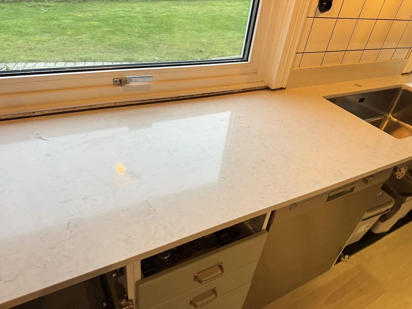 Polerad köksbänk i Silestone Snowy Ibiza i vinkel under fönstret med underlimmad rostfri diskho och vit kakelvägg i ett kök i Halmstad.