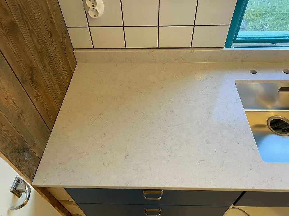 Närbild av polerad Silestone Snowy Ibiza som köksbänkskiva i Höganäs, med ljus kvartskomposit, rostfri ho och trävägg intill.