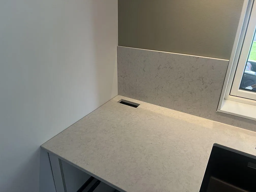 Hörn av köksbänkskiva i Silestone Snowy Ibiza, polerad komposit, med stänkskydd och synlig diskho vid fönster i Hovmantorp.