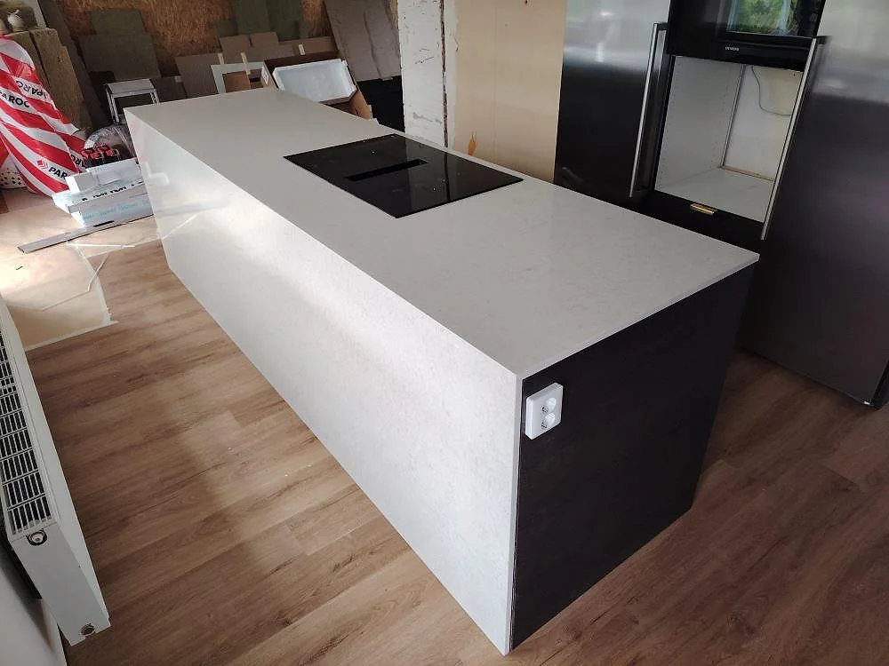 Köksö i polerad Silestone Snowy Ibiza, vit kvartskomposit, med svart häll och neddragen sida i ett kök under montering i Järna.