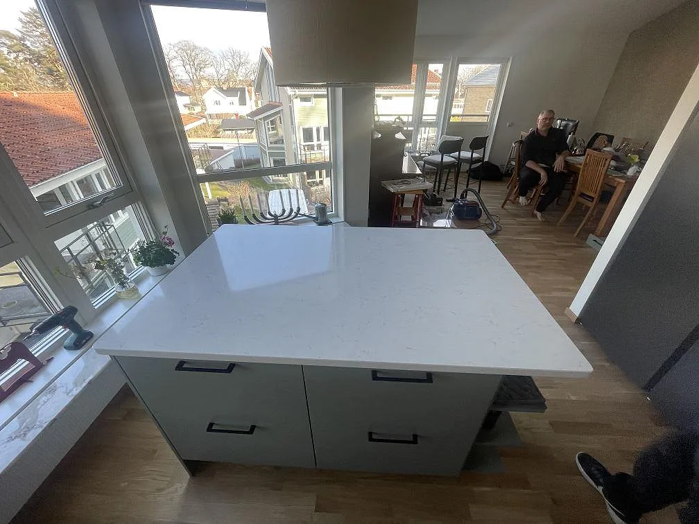 Köksö i vit Silestone Snowy Ibiza, polerad, mot dämpat gröna luckor och fönster i full höjd i ett kök i Jönköping.