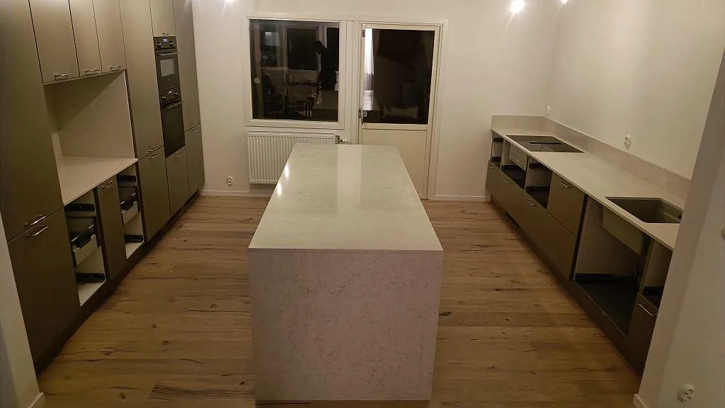 Köksbänkskiva i vit Silestone Snowy Ibiza i ett kök i Kivik, med fristående köksö i mitten och en lång bänkrad längs väggen, fotograferad under montering.