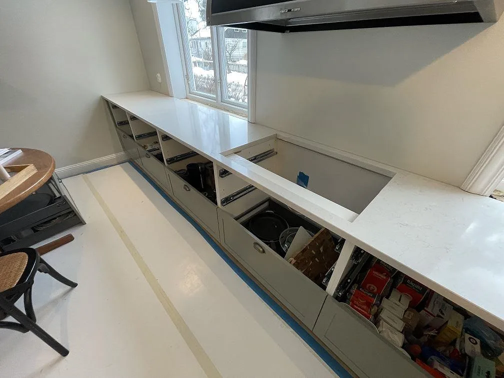 Lång rak bänkskiva i polerad vit Silestone Snowy Ibiza längs fönsterväggen, med öppna köksstommar under montering i ett kök i Lidingö.
