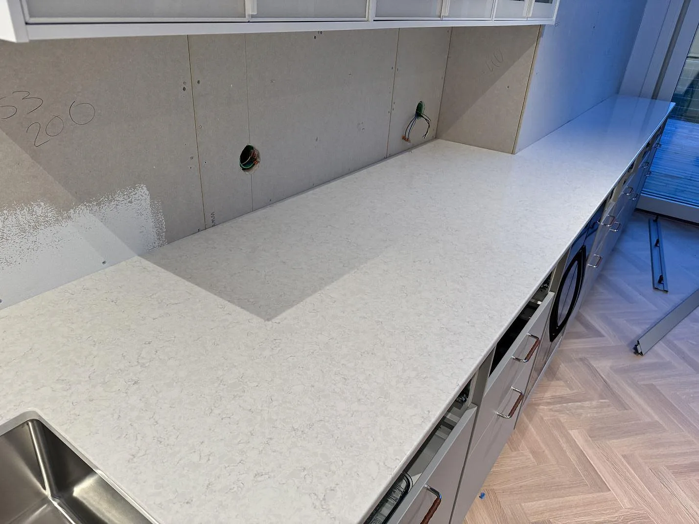 Lång köksbänkskiva i polerad Silestone Snowy Ibiza längs vägg med öppna stommar och pågående montering i Lindome.