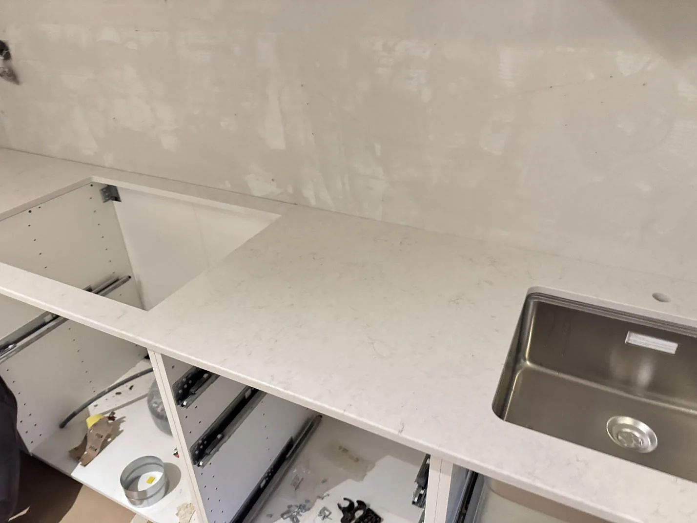 Polerad Snowy Ibiza-bänkskiva i ett kök i Malmö, med underlimmad rostfri diskho till höger och ett öppet hällurtag till vänster, fotad mot bar gipsvägg under montering.