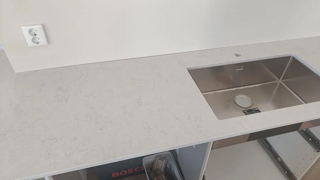 Detalj av polerad Silestone Snowy Ibiza i ljus kvartskomposit runt en rostfri diskho på köksbänkskivan i Osby.