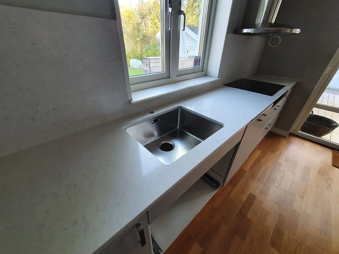 Köksbänkskiva i Silestone Snowy Ibiza, polerad, med underlimmad rostfri diskho och induktionshäll vid fönstret i ett kök i Saltsjö-Boo.