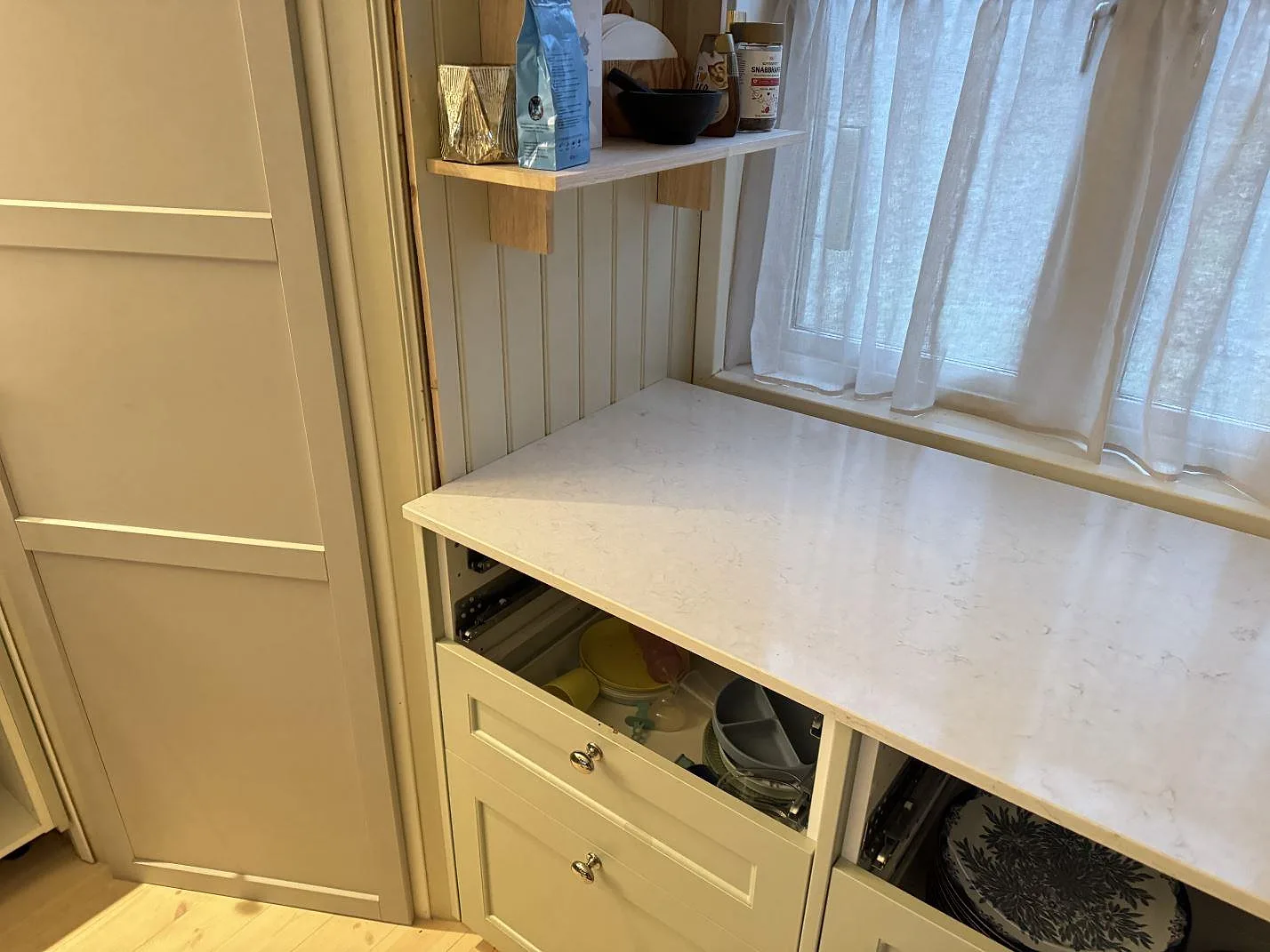 Polerad Silestone Snowy Ibiza bänkskiva i komposit längs fönstervägg med öppna underskåp i Sjuntorp.