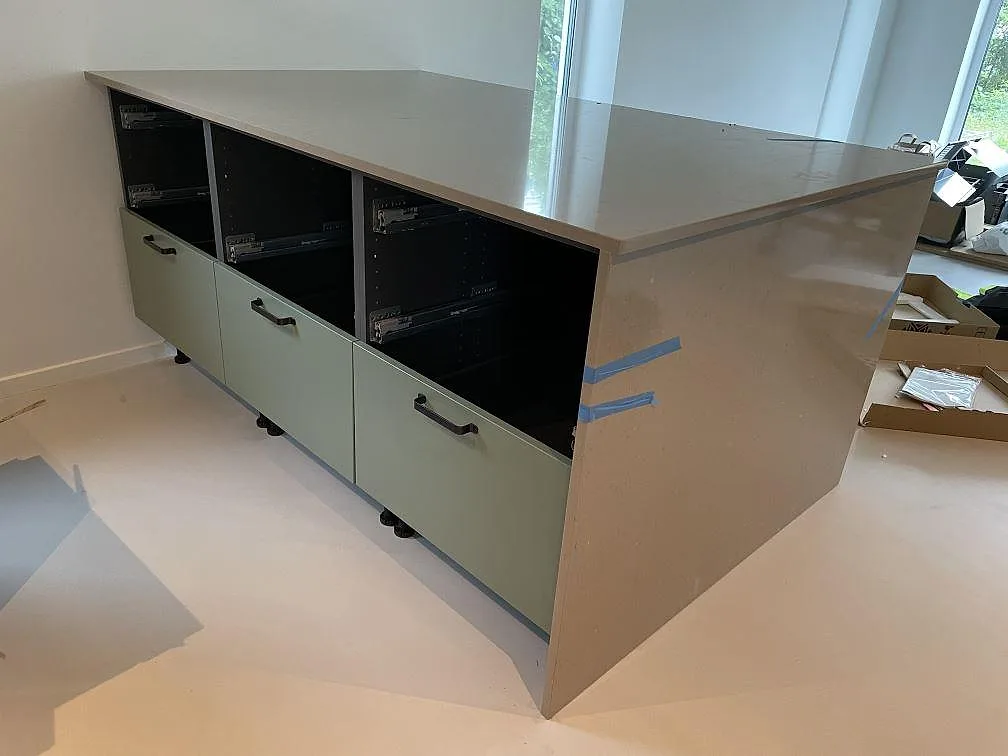 Köksö klädd med Silestone Snowy Ibiza i ljus komposit, med grågröna fronter och öppna stommar i ett kök i Skurup under montering.