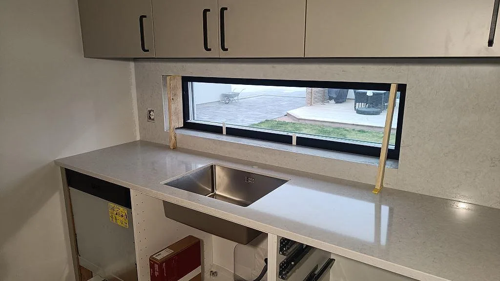 Köksbänkskiva i polerad Silestone Snowy Ibiza med rostfri diskho under fönstret och stenklädd vägg i Skurup.