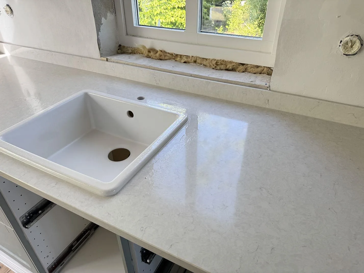 Polerad köksbänkskiva i Silestone Snowy Ibiza, ljus kvartskomposit med diskhoområde, låg stänkkant och ofärdig fönsternisch i Smygehamn.