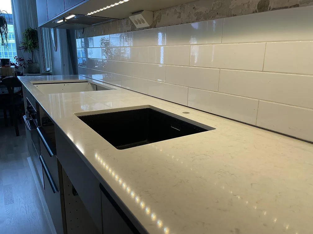 Närbild på polerad Silestone Snowy Ibiza köksbänkskiva i kvartskomposit med svart diskho, ljus ådring och kaklat stänkskydd i Stockholm.