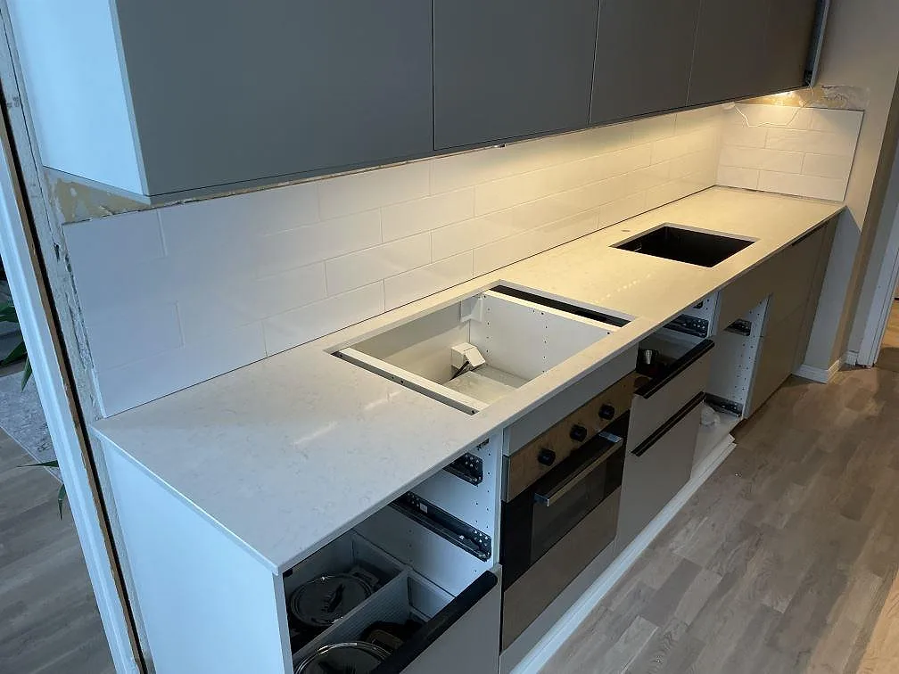 Lång köksbänkskiva i polerad Silestone Snowy Ibiza med hällöppning, svart diskho och grå köksstommar under montering i Stockholm.