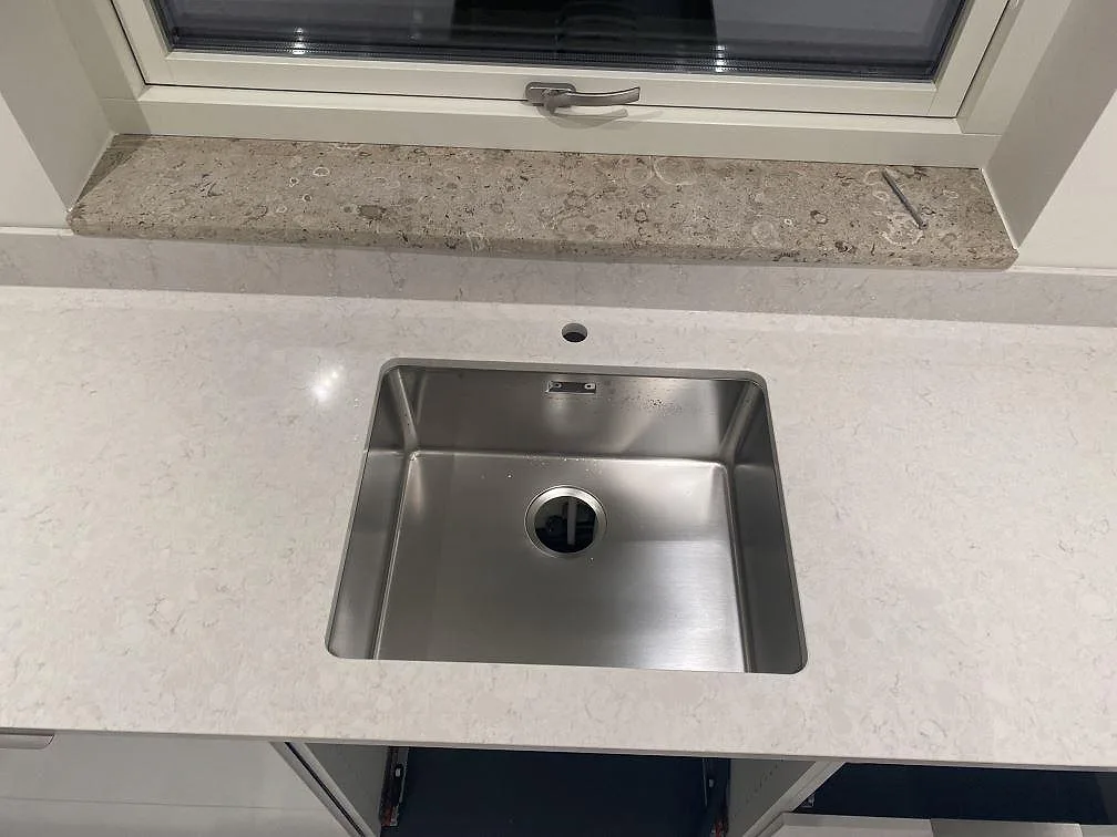 Närbild av rostfri diskho i köksbänkskiva av polerad Silestone Snowy Ibiza, ljus kvartskomposit med grå ådring, i Strandbaden.
