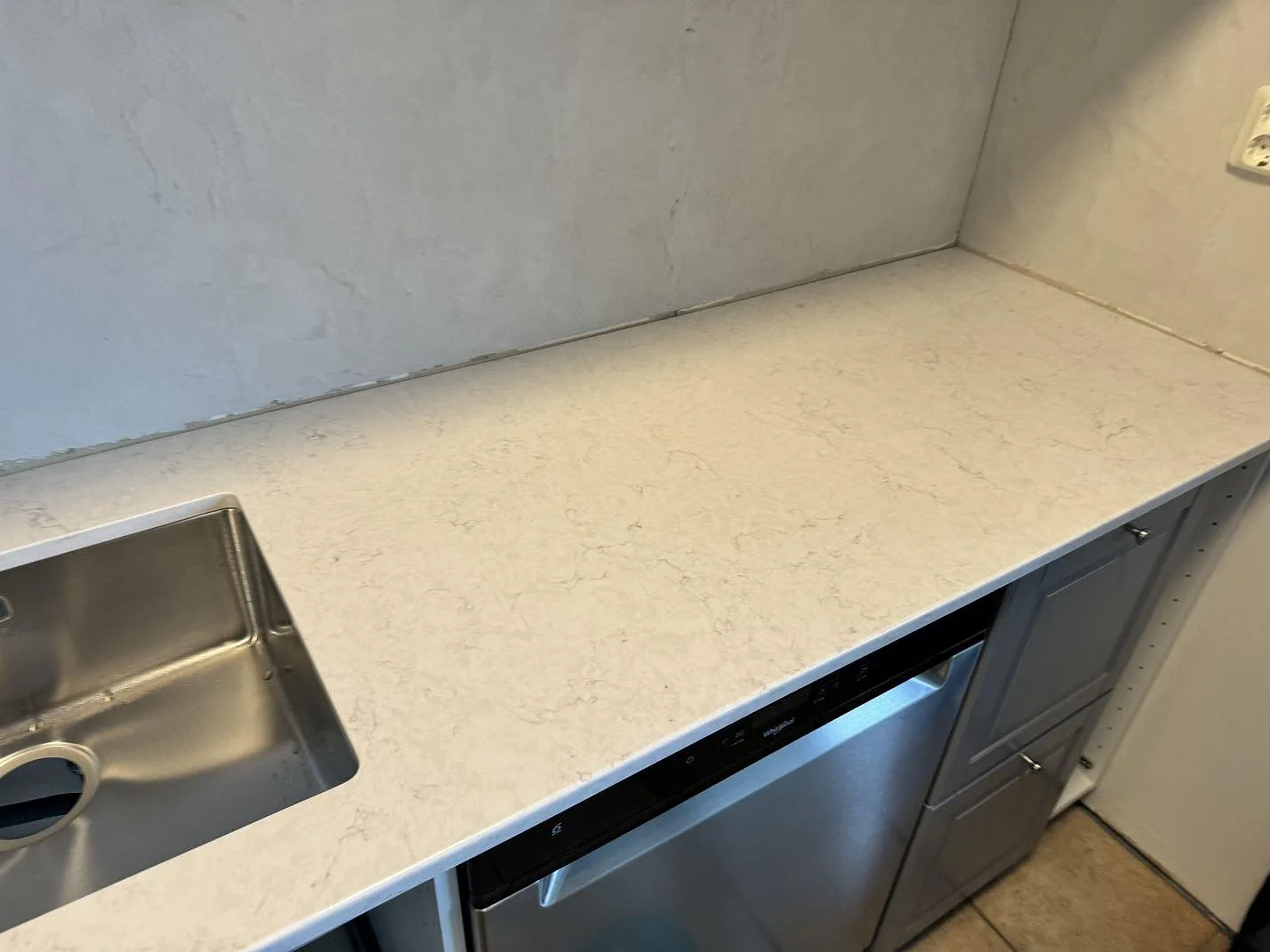 Lång köksbänkskiva i polerad Silestone Snowy Ibiza kvartskomposit över grå köksinredning och diskmaskin under montering i Svedala.