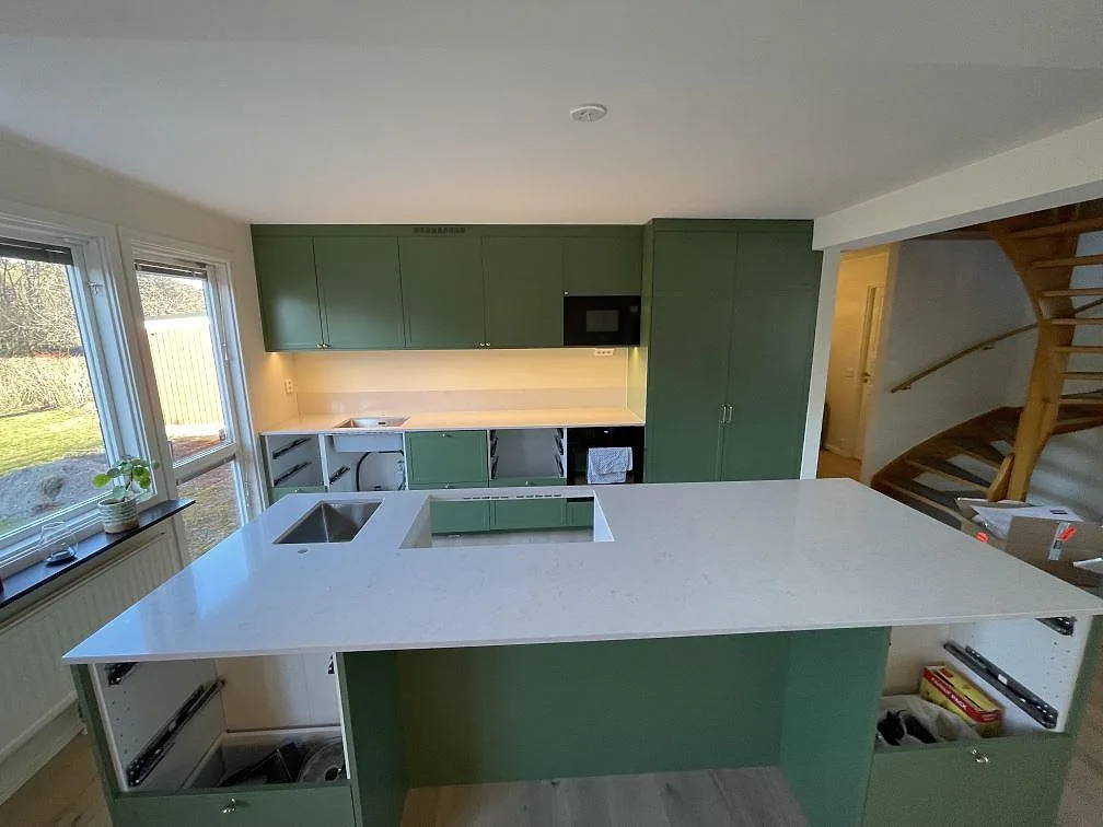 Köksö i polerad Silestone Snowy Ibiza med rostfri diskho, hällurtag och öppna gröna stommar i ett kök under montering i Täby.