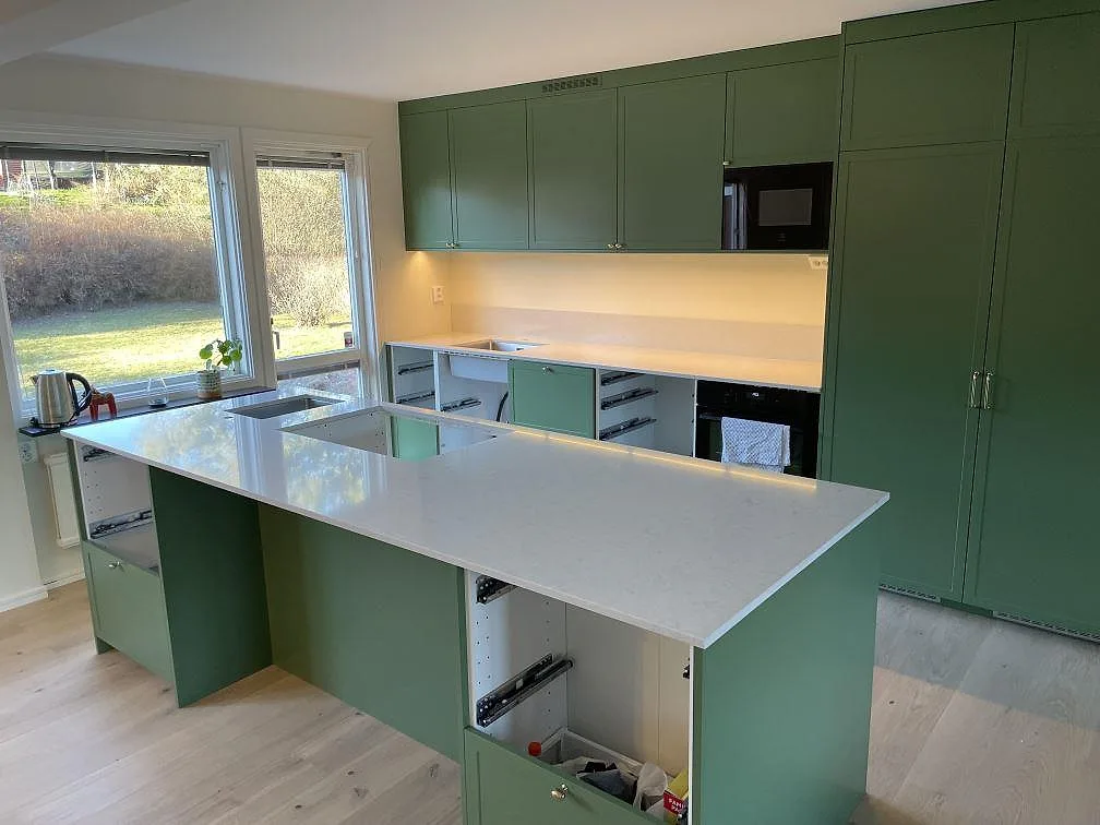 Polerad köksbänkskiva i vit Silestone Snowy Ibiza, 20 mm, med stor köksö och låg stänkkant i ett grönt kök under montering i Täby.