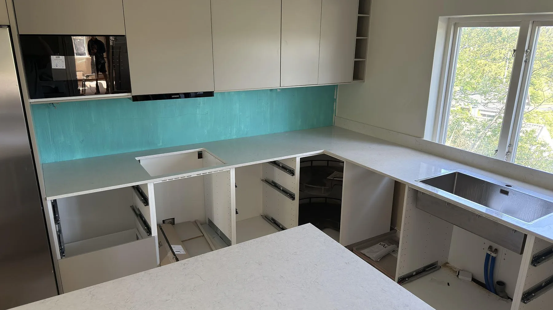 Översikt av ett U-format kök i Täby med vita bänkskivor i Silestone Snowy Ibiza, mintgrönt glasstänkskydd och öppna stommar under montering.