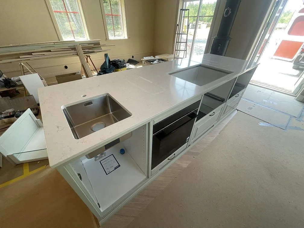 Stor köksö med polerad Silestone Snowy Ibiza, rostfri ho och hällurtagning i ett ljust kök under montering i Trosa.
