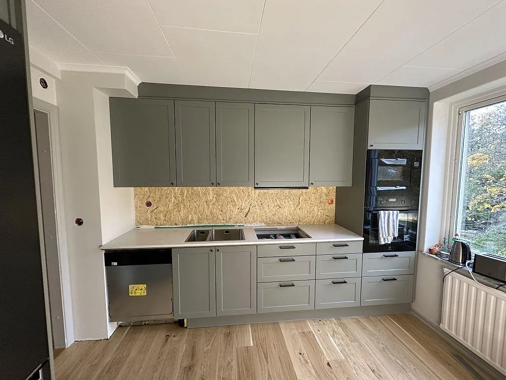 Kök i Uddevalla med salviagröna luckor, vit Silestone Snowy Ibiza som bänkskiva och temporär OSB-skiva bakom arbetsytan.
