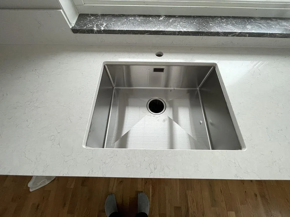 Underlimmad rostfri diskho infälld i en vit polerad Silestone Snowy Ibiza-bänkskiva, med mörk stenfönsterbänk i bakgrunden, i ett kök i Västerås.