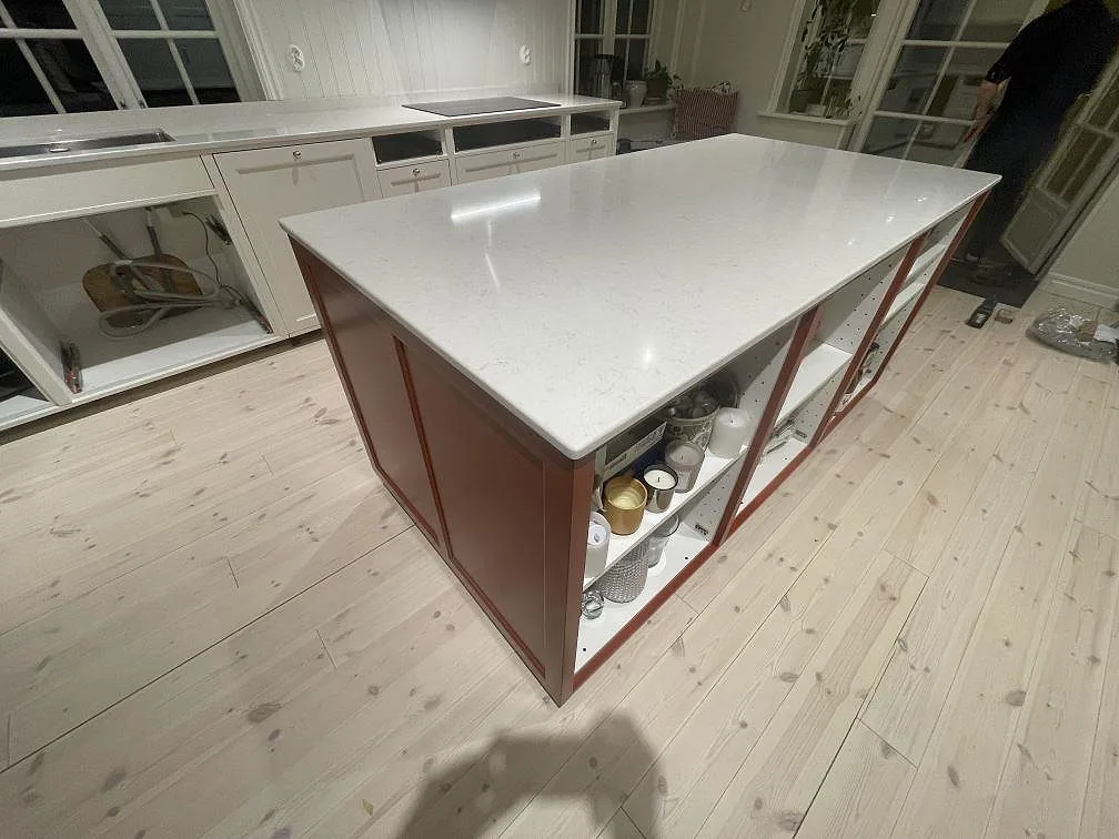 Köksö i polerad Silestone Snowy Ibiza sedd från sidan med öppna displayhyllor och bänkparti med induktionshäll i bakgrunden, Vallentuna.