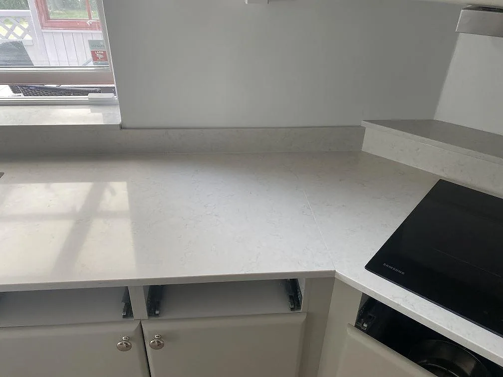 Ljus köksbänkskiva i polerad Silestone Snowy Ibiza vid fönster och svart häll, med öppna underskåp i Ystad.