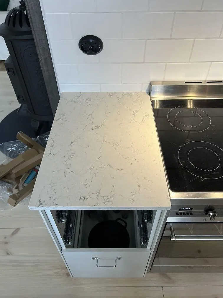 Köksbänkskiva i Silestone White Arabesque, polerad kvartskomposit, bredvid spis och öppna vita skåpsstommar i Villands Vånga.