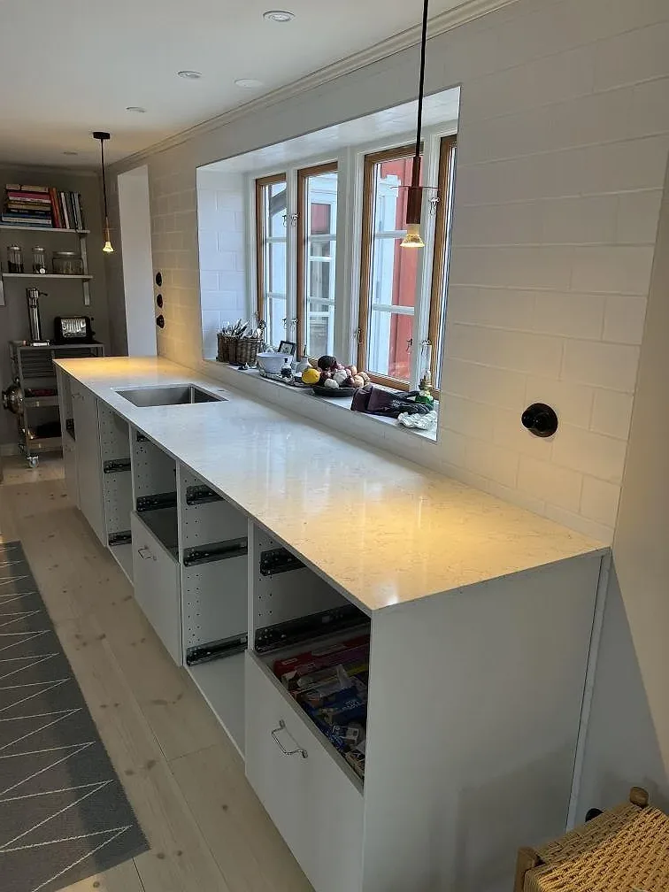 Lång köksbänkskiva i polerad Silestone White Arabesque med vit kakelvägg, fönsterljus och öppna skåpsstommar i Villands Vånga.