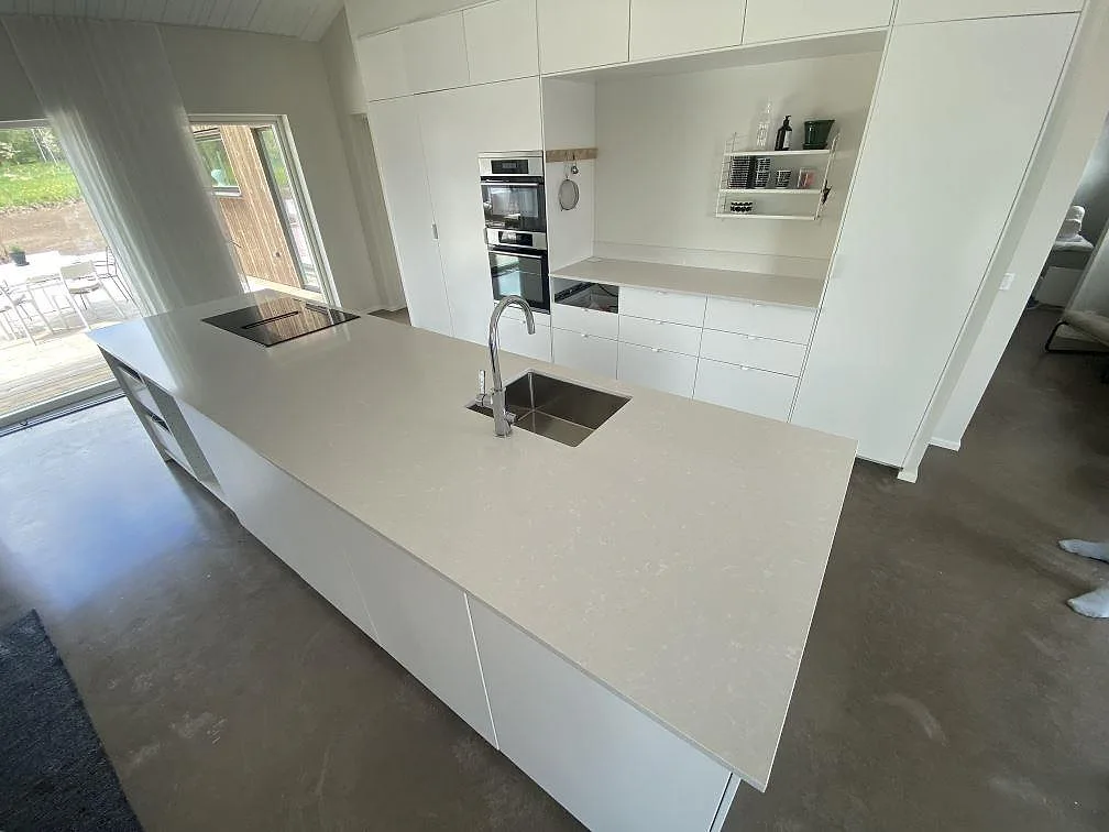 Köksbänkskiva i Silestone Yukon, polerad greige kvartskomposit, på en stor köksö med underlimmad rostfri diskho och induktionshäll i ett ljust kök i Sävsjö.