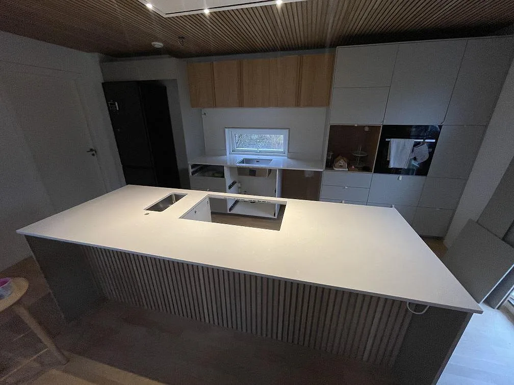 Köksbänkskiva i polerad Silestone Yukon, ljus kvartskomposit, på en stor köksö med öppning för häll och synlig rostfri diskho i Tyresö.