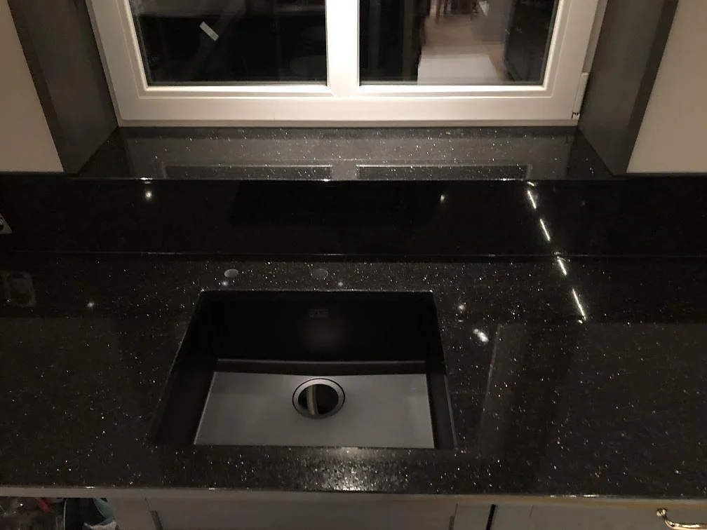Underlimmad diskho i en bänkskiva av svart Star Galaxy granit vid ett fönster, med borrade kranhål och glittrande yta, i ett kök i Alingsås.