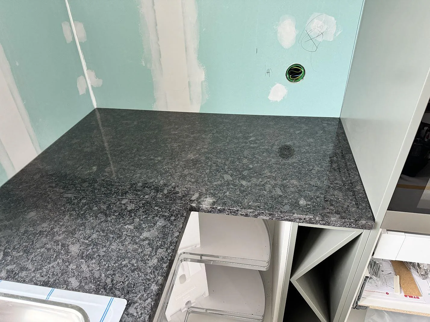 Hörnparti av köksbänkskiva i polerad Steel Grey granit med öppen förvaring under montering i Älmestad.
