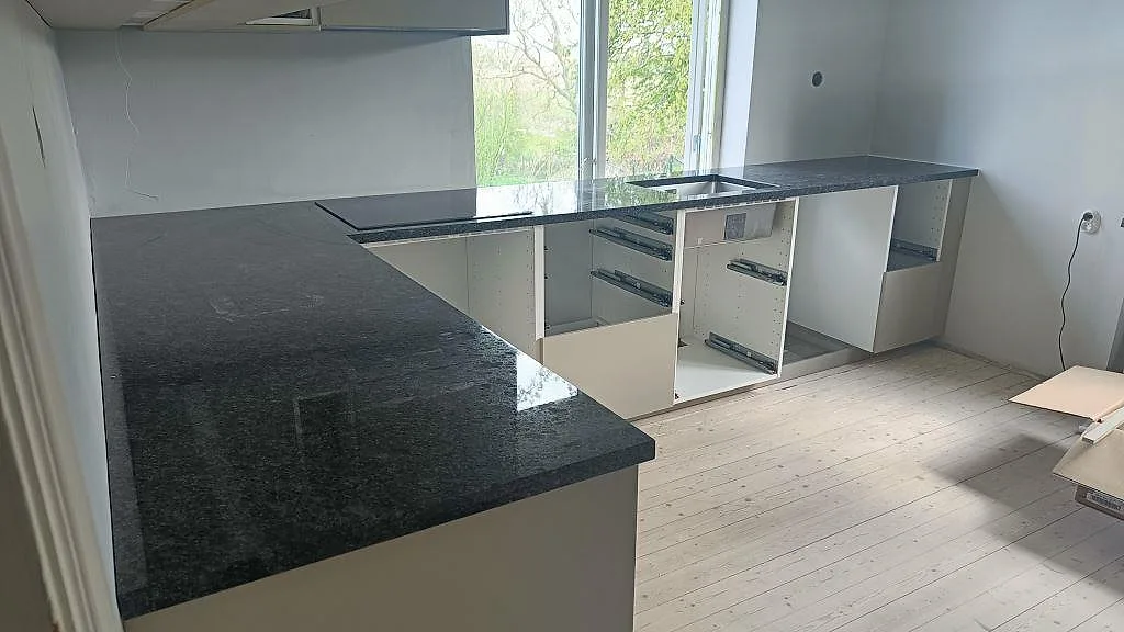 Vinklat kök i polerad Steel Grey granit i Anderslöv, med infälld induktionshäll och underlimmad rostfri diskho, fotograferat under montering.