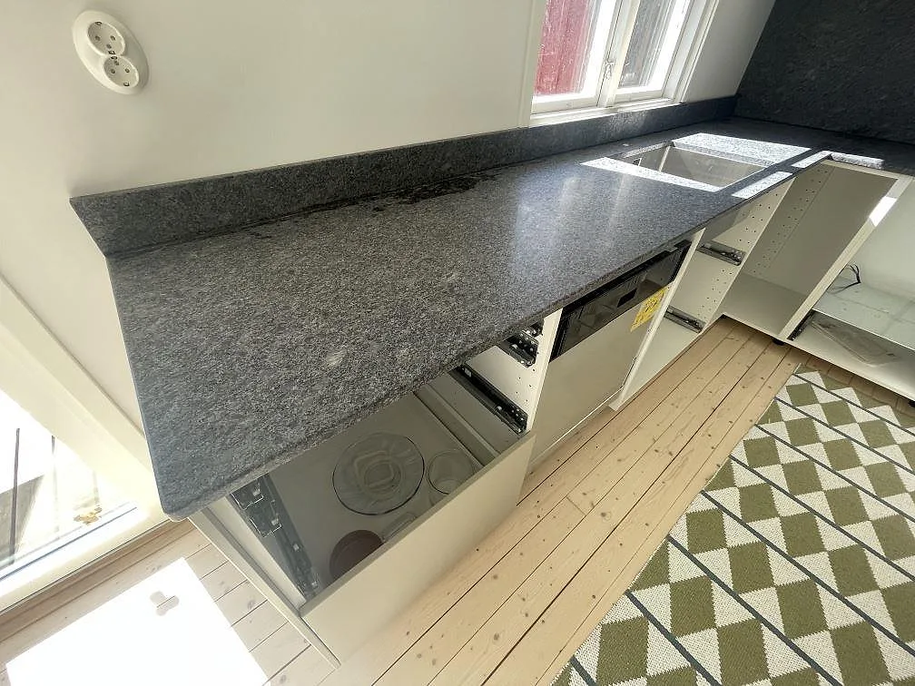 Närbild på köksbänkskiva i matt Steel Grey granit med hoöppning och låg stänkkant vid fönster i Grisslehamn.