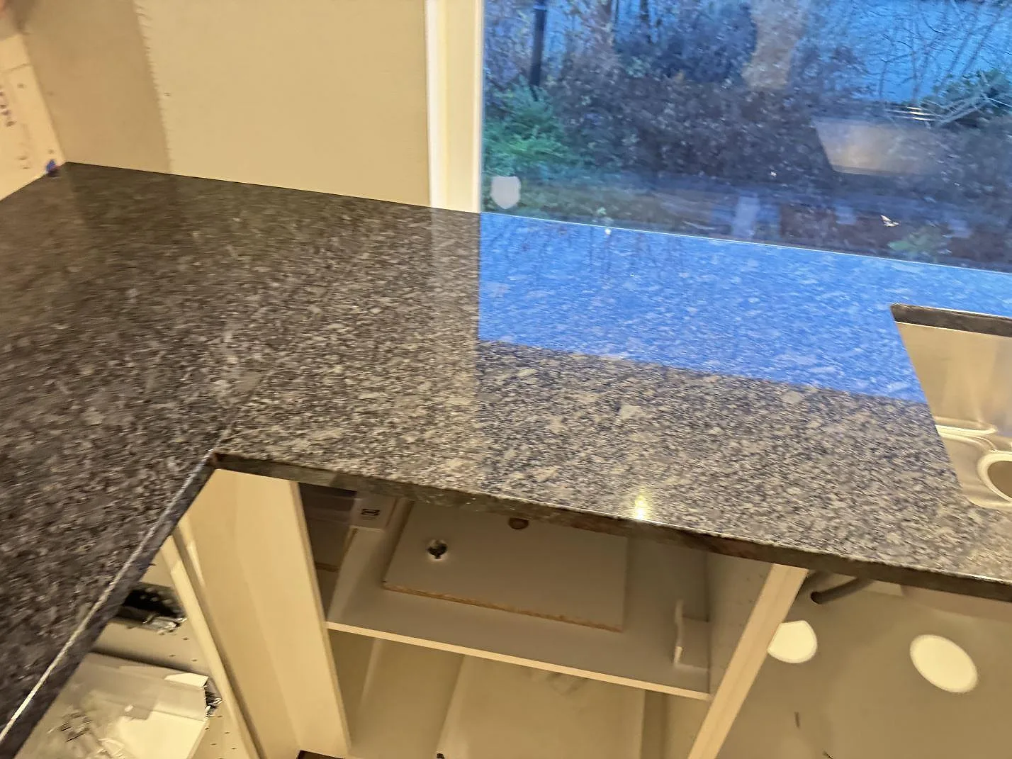 Lång bänkyta i polerad Steel Grey-granit med diskhoöppning och fönsterreflektion i ett kök i Haverdal.
