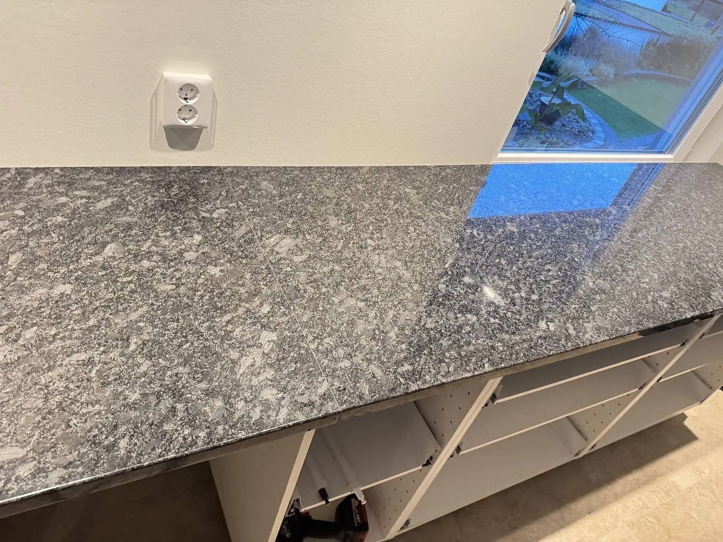 Lång köksbänkskiva i polerad Steel Grey-granit med eluttag på väggen och öppna vita stommar i Haverdal.