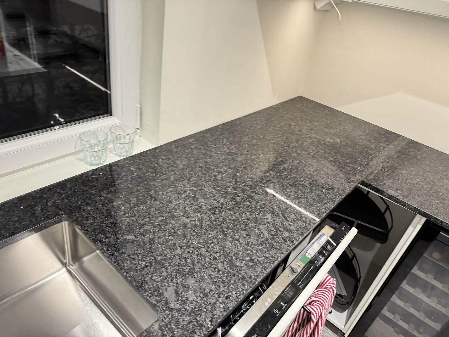 Köksbänkskiva i Steel Grey-granit med infälld induktionshäll och rostfri diskho i ett hörn i ett kök i Kävlinge.