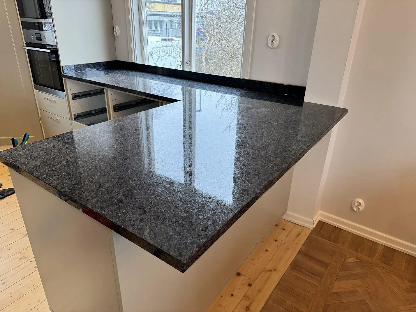 Blank halvö i Steel Grey-granit med fönsterreflexer och öppen kökslösning i Mölndal.