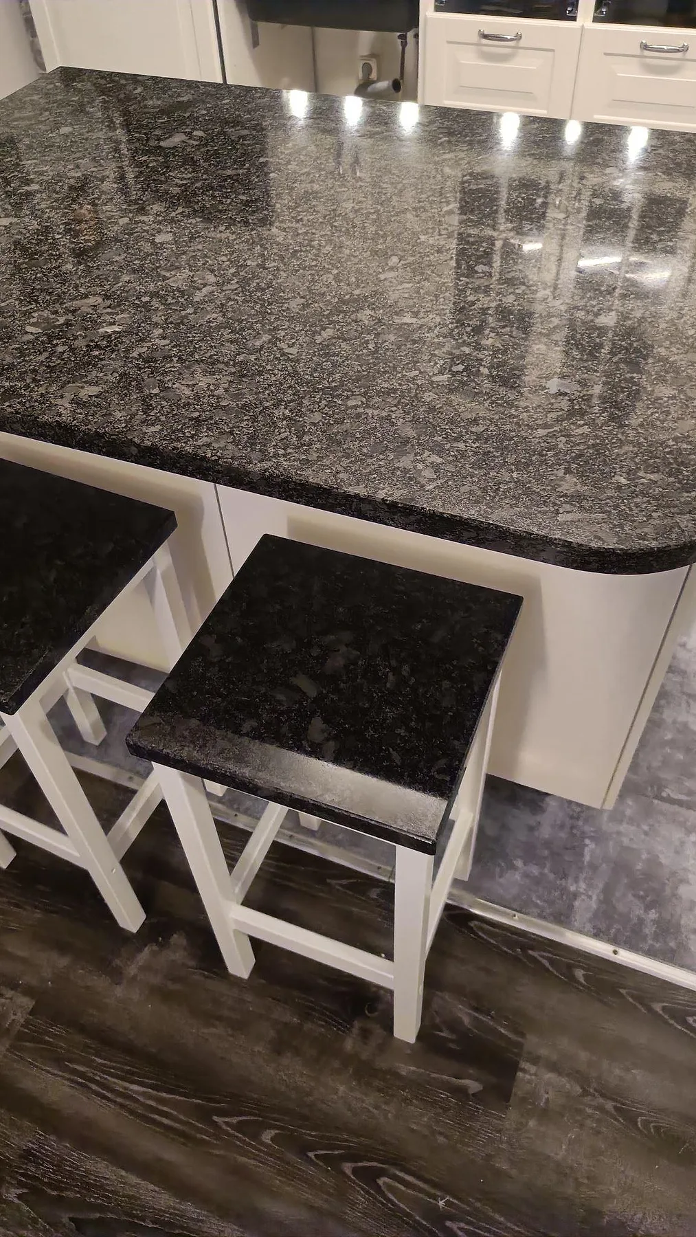 Närbild på köksö i polerad Steel Grey granit med mörka barstolssitsar och vita underskåp i Oxie.