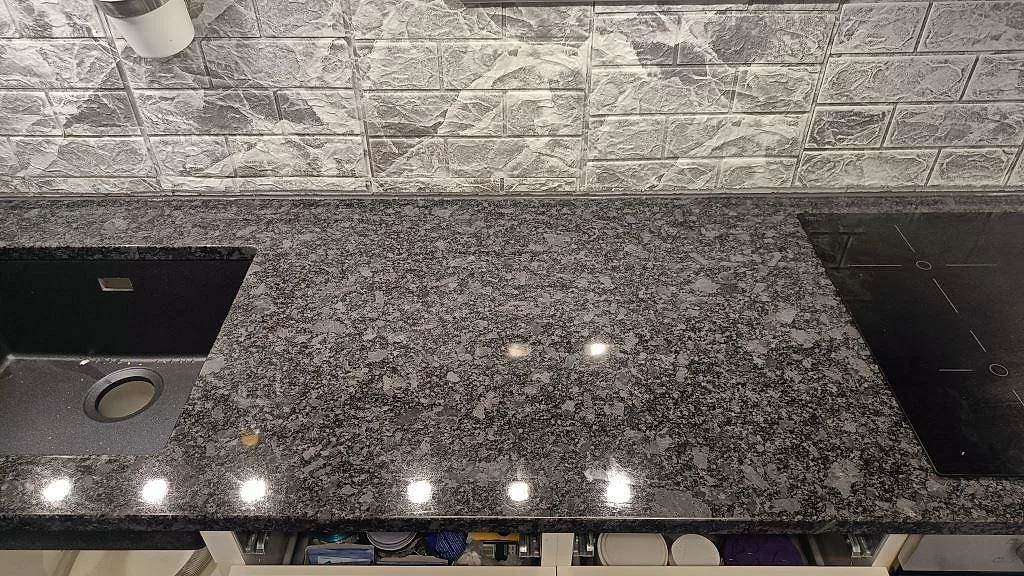 Köksbänkskiva i polerad Steel Grey granit mellan mörk diskho och infälld häll i ett kök i Oxie.