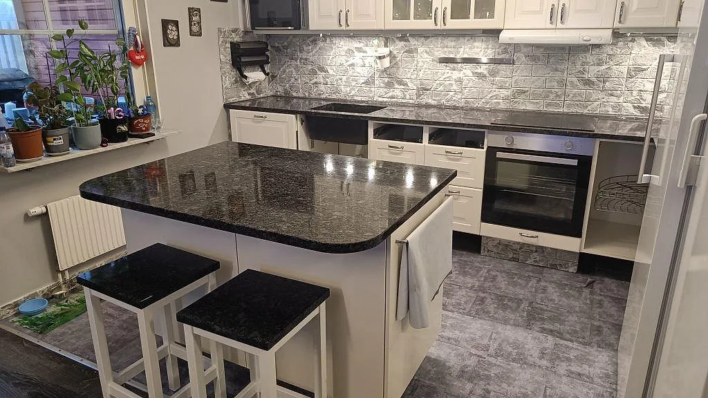 Köksbänkskiva och köksö i polerad Steel Grey granit med vita köksluckor, barstolar, diskho och häll i Oxie.