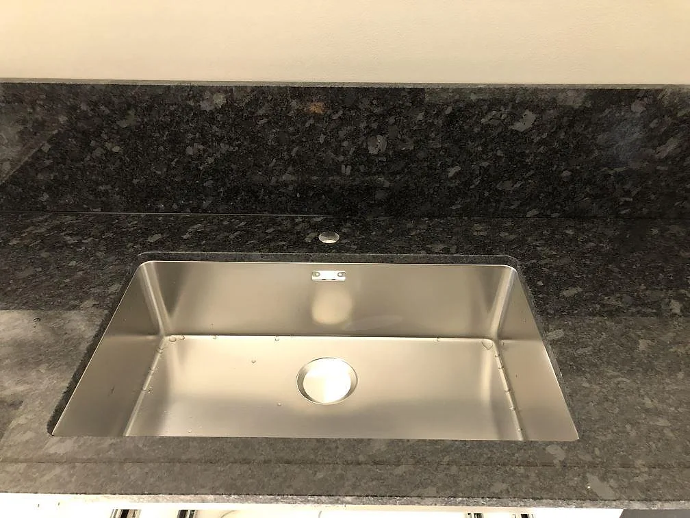 Underlimmad rostfri diskho infälld i en köksbänkskiva i Steel Grey granit med uppvikt stänkkant i ett kök i Södra Rörum.