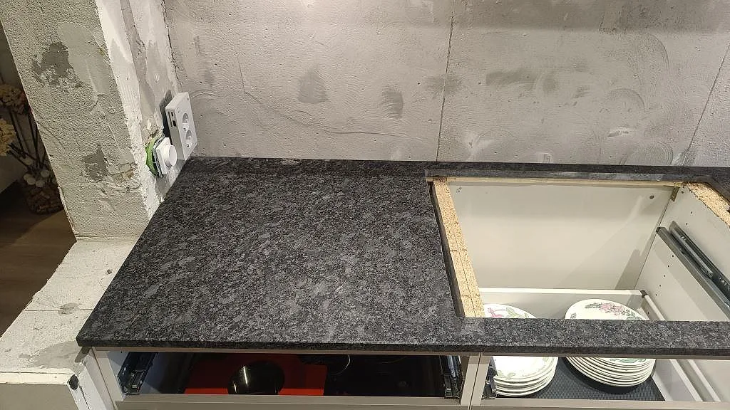 Installerad Steel Grey granitbänkskiva med öppna lådfronter och rå väggyta i ett kök i Staffanstorp.