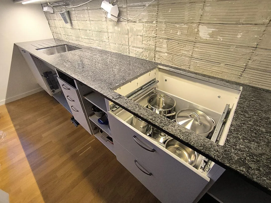 Lång köksbänkskiva i polerad Steel Grey granit med diskho och hällöppning framför öppen köksstomme i Sundbyberg.